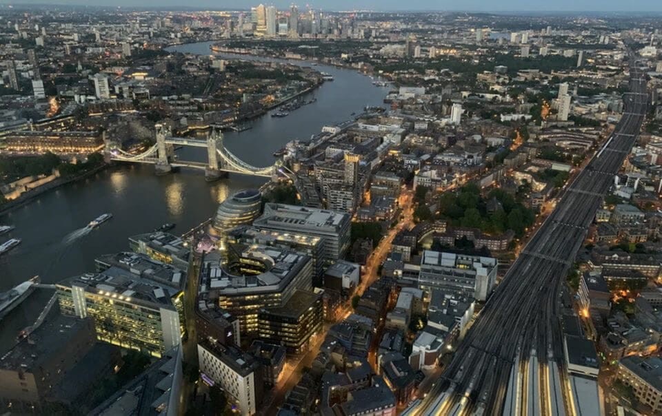 Aerial view of London at dusk, showcasing the Tower Bridge over the River Thames, with the glow of city lights starting to illuminate the streets and buildings, highlighting the intricate urban planning and architectural diversity.