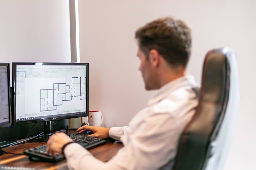 Professional architect working on a building plan in CAD software on a dual monitor setup, with focus on the design process and precision in architectural planning, in a well-lit modern office environment.