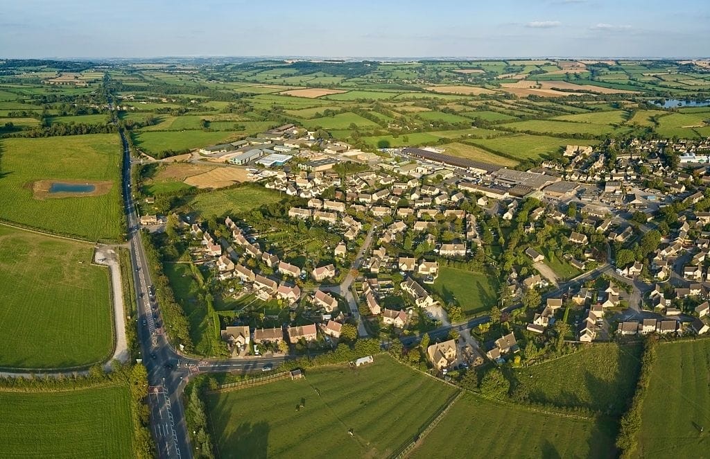 Breathtaking aerial view of a quaint English village at sunset with lush green fields, cosy clustered houses, and a clear main road, showcasing rural beauty and village life in the UK.