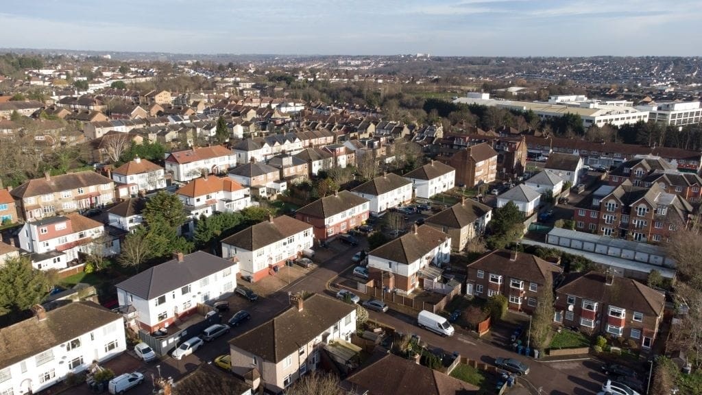 Aerial view of a suburban neighborhood in the UK showcasing semi-detached houses with varying facades and tiled roofs, interspersed with roads and vehicles, on a sunny day with a clear sky.