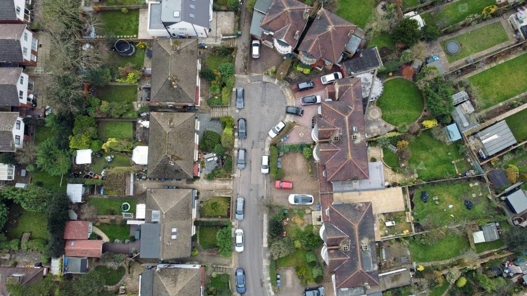 Direct overhead view of a residential area in the UK with detailed gardens, a variety of detached and semi-detached homes, and a winding road lined with parked cars, depicting typical suburban planning and architecture.