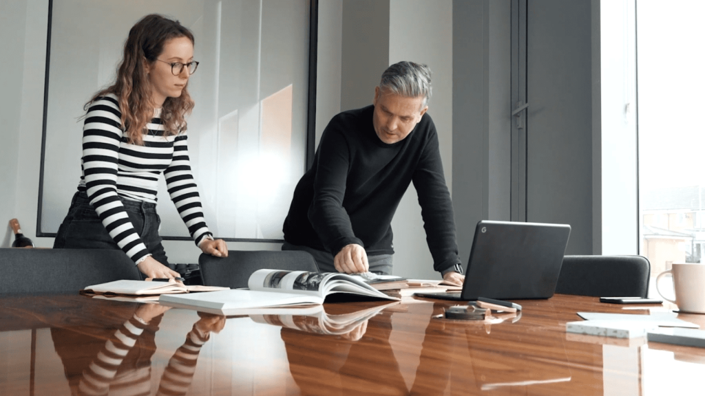 Two architects reviewing blueprints and architectural plans at a conference table in a well-lit office, with a laptop and coffee mug in view.