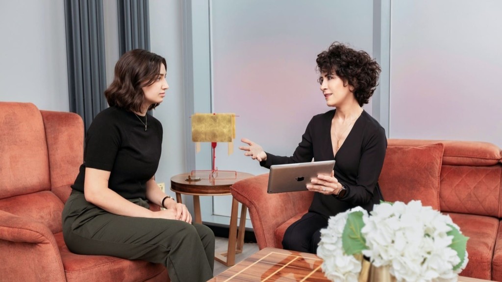 Two women sitting on burnt orange couches in a modern office, engaged in a professional discussion about architectural design. One woman, holding a tablet, is explaining a concept with hand gestures, demonstrating her specialised knowledge in the field while the other listens attentively. A decorative lamp and a table with a flower arrangement emphasise the stylish interior design.