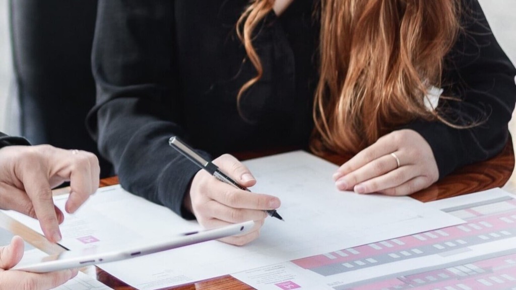 Two architects collaborating over detailed building plans at a wooden table, with one pointing to a design on a digital tablet and the other holding a pen, indicating collaboration while liaising with the Local Planning Authority (LPA). The table is covered with architectural drawings that reflect the importance of teamwork in planning.