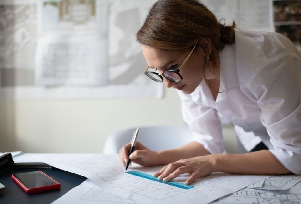 Concentrated female architect analysing floor plans on a desk with a smartphone and stationery aside, in a well-lit workspace with project blueprints in the background.