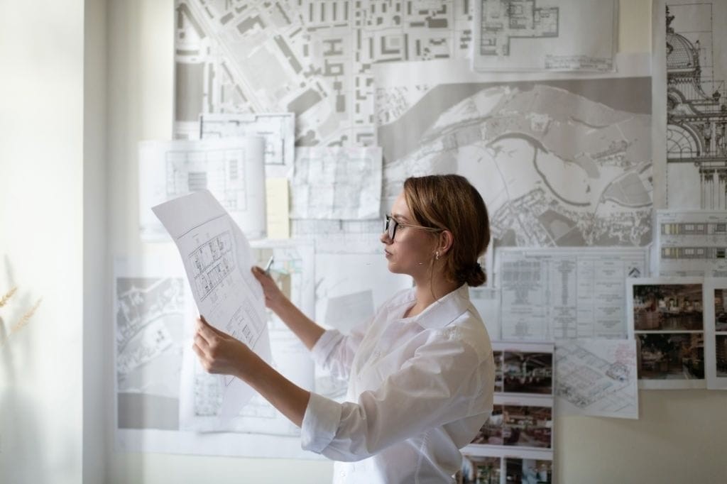 Female architect reviewing architectural plans in a bright office with wall-mounted urban development blueprints in the background.