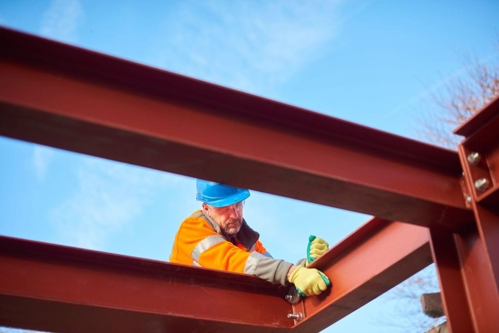 Construction worker in high-visibility jacket and safety helmet securing steel beams on a construction site against a clear blue sky, representing industrial development and construction safety standards.