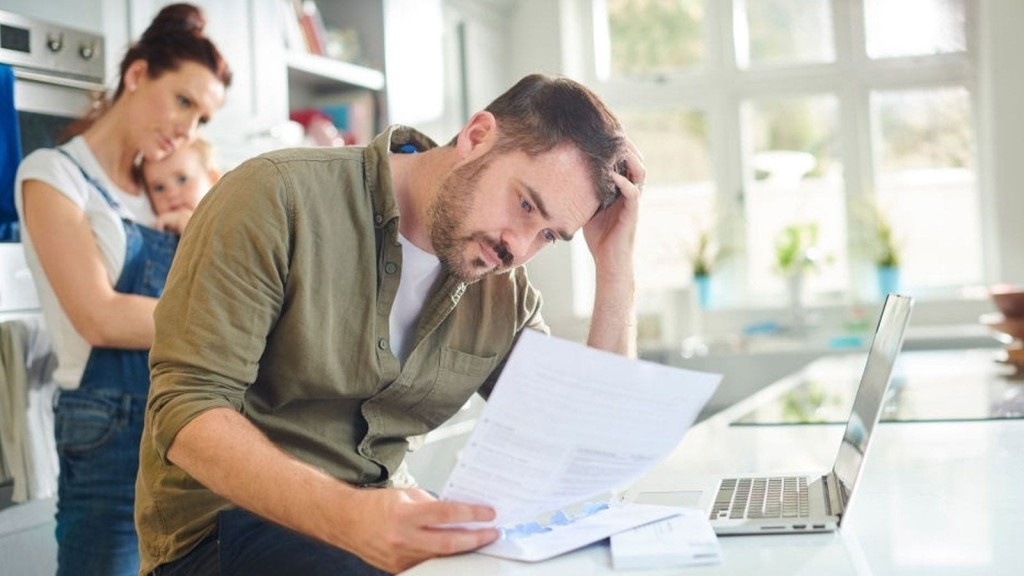 Concerned man reviewing financial documents at kitchen counter with laptop while woman holding a child looks on, depicting family financial planning for construction works.