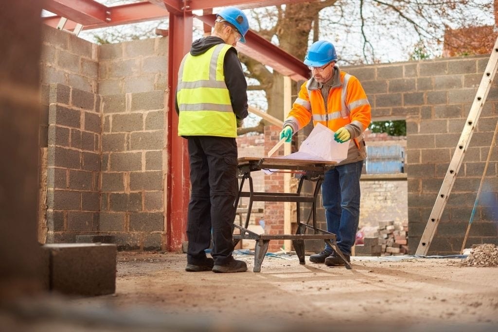 Two construction workers in safety gear examining blueprints at a residential building site with steel beams and cinder block walls in the background.