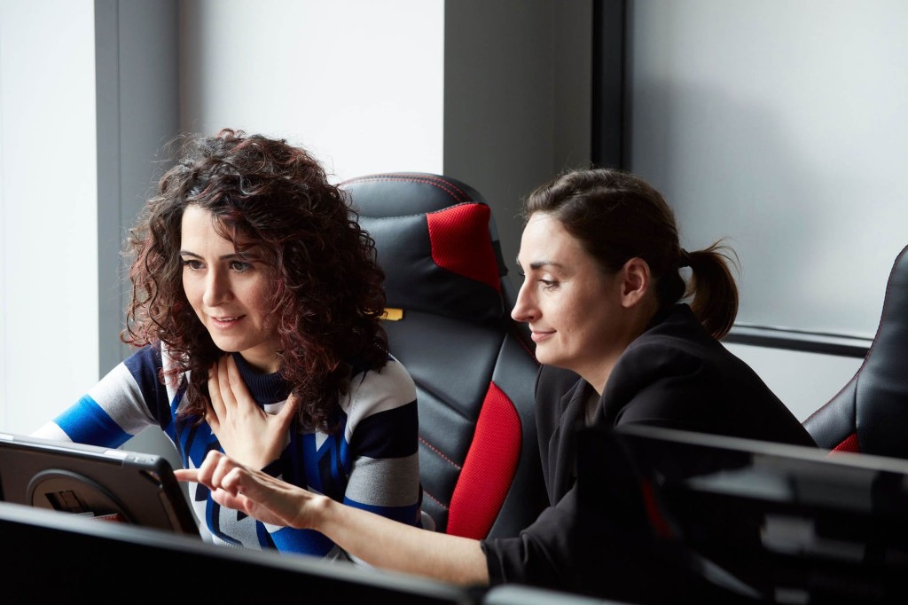 Two professional women collaborating at a computer in a modern office setting, focusing intently on a screen, indicative of teamwork, technology use in business, and female leadership in the workplace.