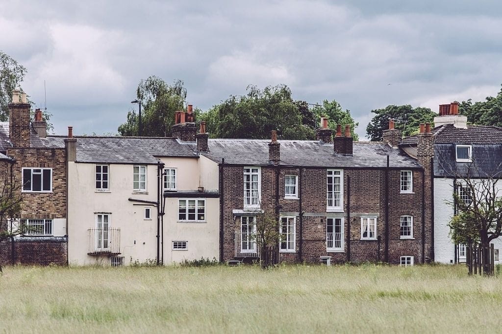 Row of traditional British terraced houses with brick facades and chimneys, set against a cloudy sky, viewed over a wild grass field.