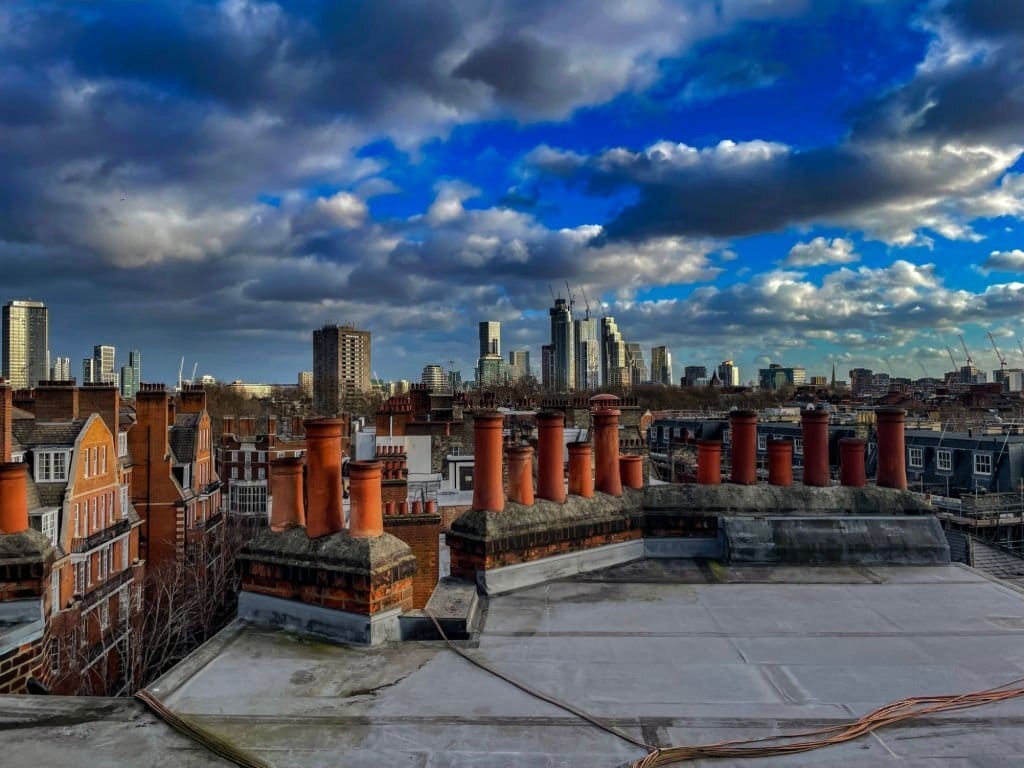 Rooftop view of London's city skyline on a cloudy but blue skied day with the high rise buildings of Canary Wharf standing out in the far distant middle of the photograph