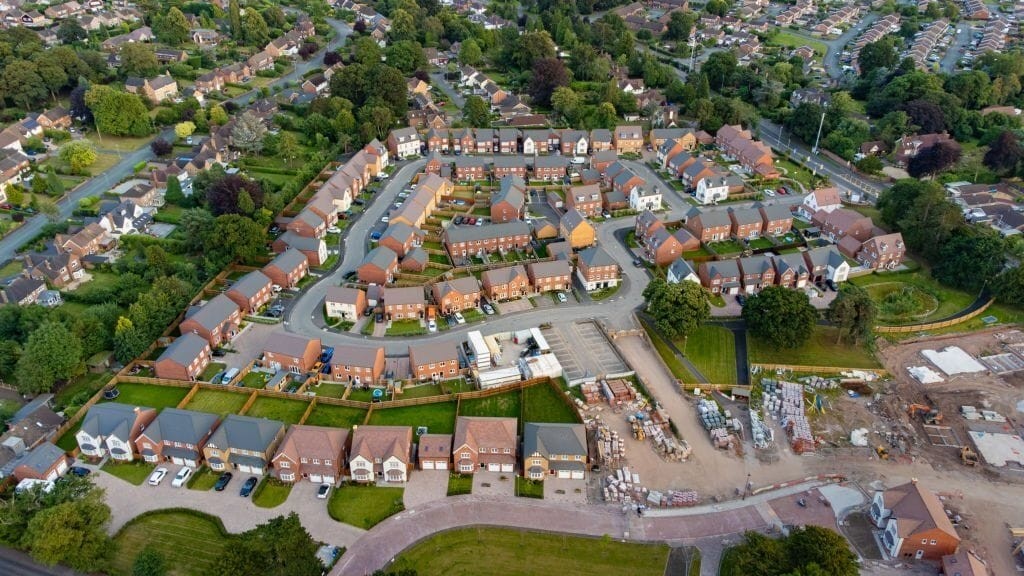 Aerial view of a suburban housing development showing a mix of completed homes with red roofs and driveways, and an ongoing construction site with building materials and machinery, surrounded by lush greenery in a peaceful neighborhood.