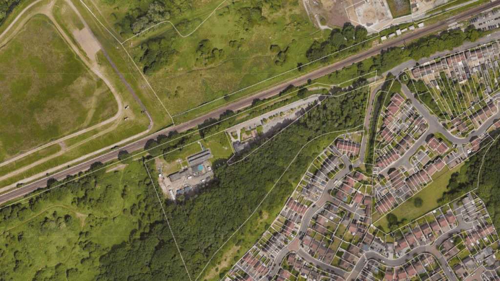 Aerial image of a suburban development next to green parkland, showcasing a juxtaposition of residential housing and natural landscape for property due diligence in urban planning. The photo features a clear demarcation with the lush park on the left, a railway line running parallel to a row of industrial facilities, and a curved residential street lined with houses on the right, illustrating the diverse land use within a compact area suitable for analysis in real estate development and urban design in North London.