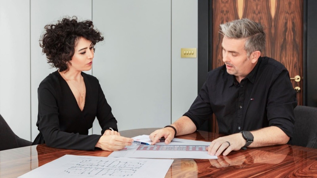 Two professional architects in a meeting, discussing over architectural blueprints and facade renderings on a polished wooden table. The male and female professionals are engaged in project planning, exemplifying a collaborative environment in architectural design and property due diligence. 