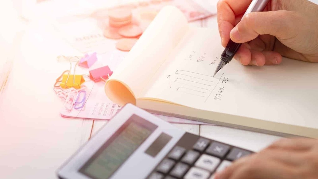 Close-up of a person's hand writing financial calculations in a notebook with a calculator, paper clips, and currency notes in the background, indicating budget planning.