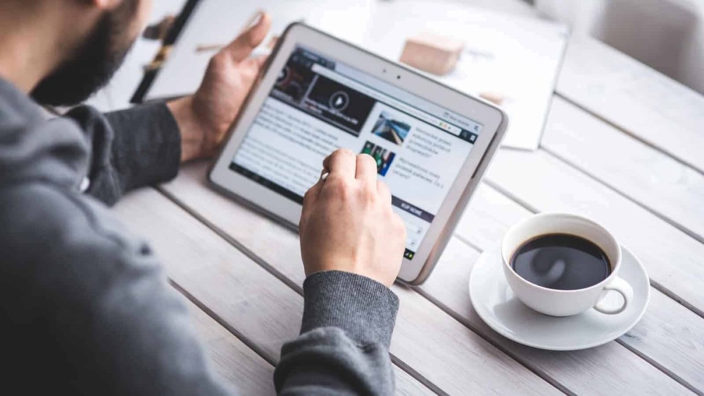 Man in gray sweater browsing real estate investment articles on a tablet with a stylus, with a fresh cup of coffee on a white wooden table, symbolising informed property investing.