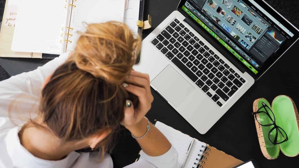 Overhead view of a stressed woman holding her head whilst looking at her laptop with photo editing software, her desk organized with an open planner, notebook, a pair of green flip-flops, and black eyeglasses, illustrating a professional multitasking in a creative workspace.