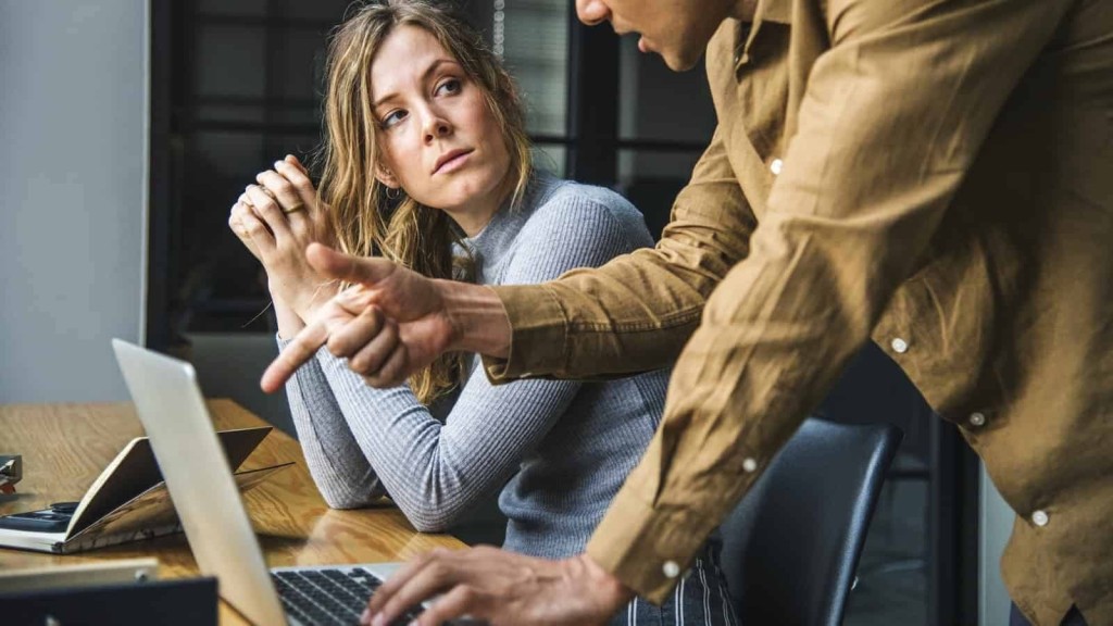 Focused businesswoman in a gray sweater looks at her colleague with concern while a male colleague in a tan shirt points something out on the laptop, highlighting a collaborative moment in a professional office environment.