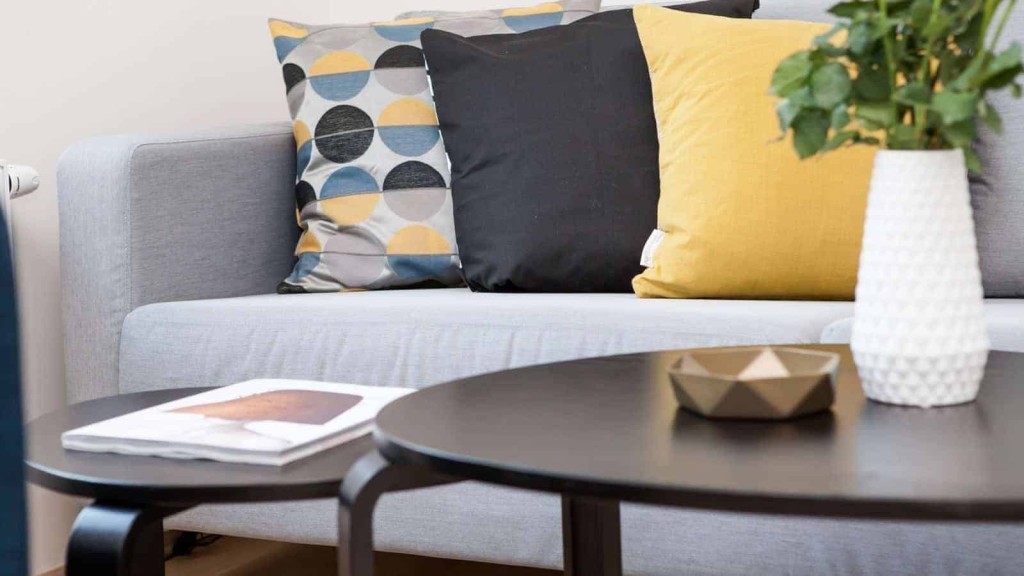 Modern living room detail with gray sofa adorned with geometric-patterned throw pillows in mustard yellow, black, and gray, alongside a white textured vase with green foliage on an end table and a round black coffee table with a geometric bowl and art book in the foreground.