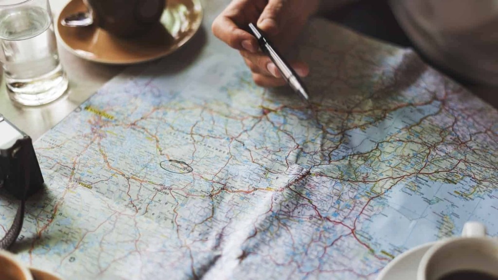 Close-up of a person's hands holding a pen over a detailed road map, with a camera and a cup of coffee on a wooden table, showcasing the search of finding the right location for their next investment.
