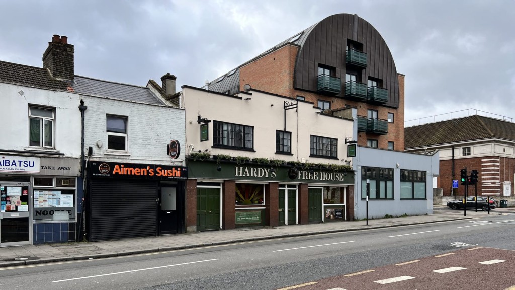 Street view of a diverse urban streetscape in Greenwich featuring a traditional pub with green facade, Hardy's Free House, alongside various local businesses including Aimen's Sushi, and a modern apartment complex with a curved architectural design and balconies.