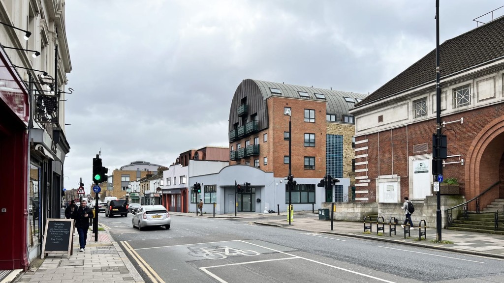 Street scene in Greenwich, London, with a dynamic skyline featuring the contrast of a modern curved apartment block with a metallic roof and traditional brick buildings including the proposed upward extension on the pub for the boutique hotel under a cloudy sky, with vehicles and pedestrians on the move.