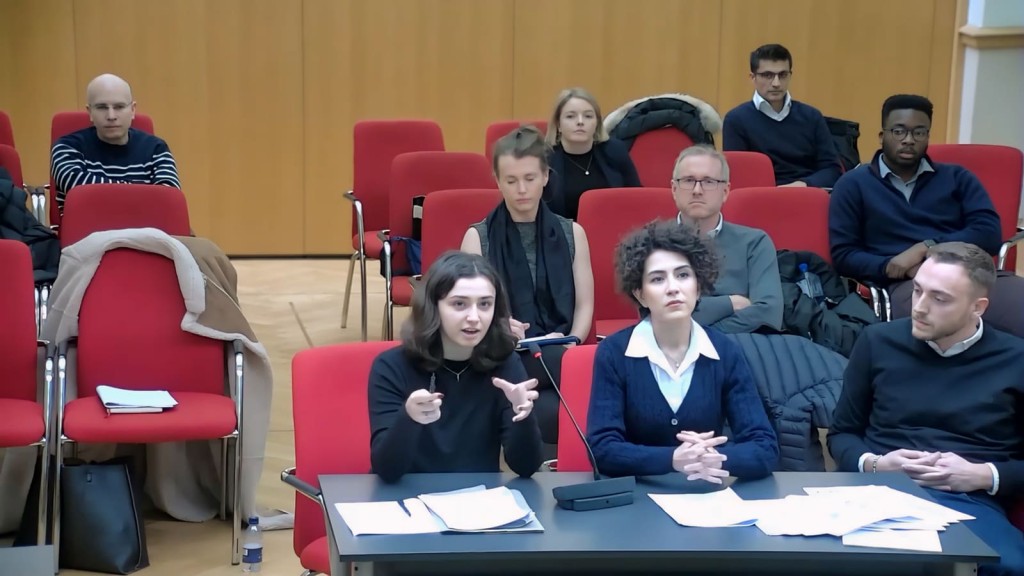 Young female planning consultant from Urbanist Architecture actively participating and asking a question during a planning committee meeting in Greenwich, with a microphone in hand, surrounded by other team members, owner and members of the public in the town hall setting.