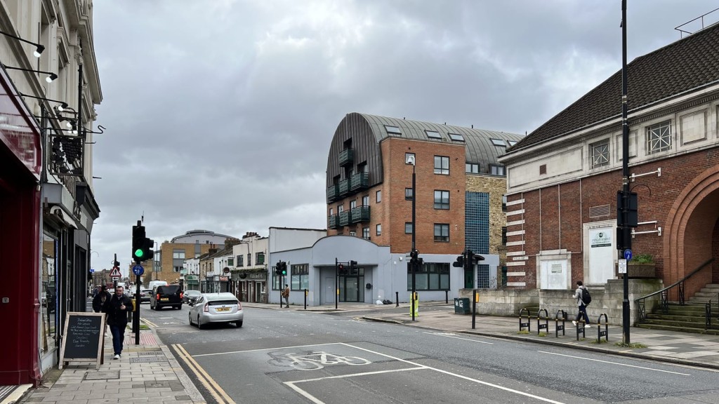Urban street view in Greenwich featuring a mix of architecture with a modern apartment block with a curved metal roof, alongside classic white and brick buildings, pedestrian walkways, and a cyclist lane under an overcast sky.