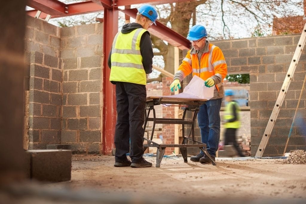 Construction site activity with two workers in high-visibility vests and safety helmets reviewing plans on a makeshift table amidst the framework of a new building, symbolising teamwork and safety in construction management.