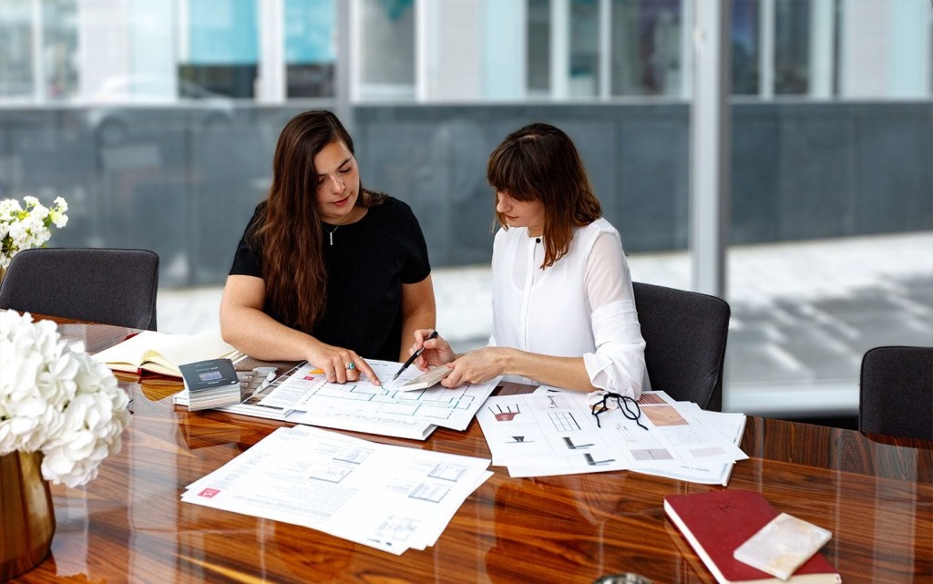 Two professional women engaged in discussion over architectural plans spread out on a polished wooden table, with fabric samples, indicating a collaborative design session in a modern office setting with natural light.