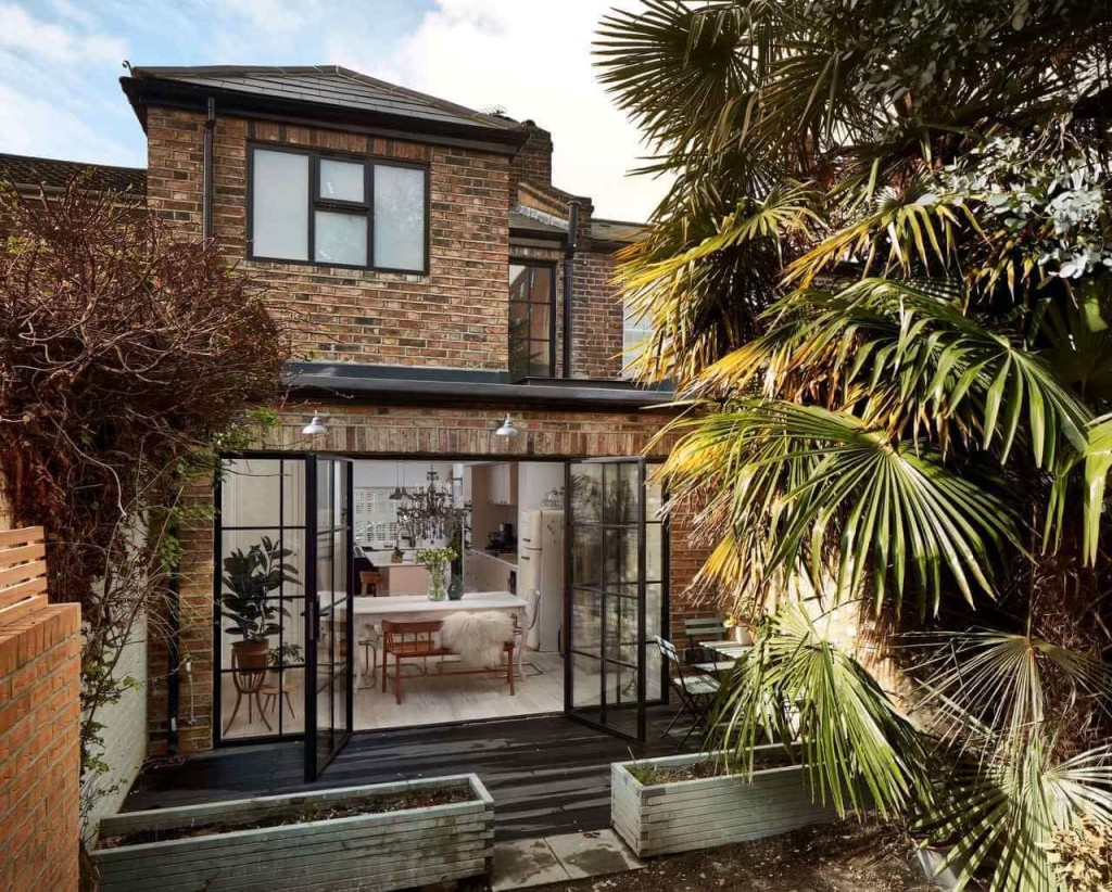Chic rear house extension with black steel-framed glass doors opening into a bright dining area with a classic design, nestled in a lush garden with tropical plants, showcasing a blend of traditional brick architecture and modern glass elements.