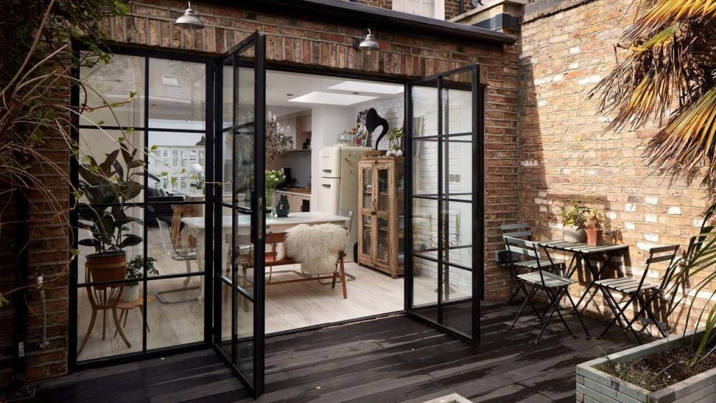 View from a cosy urban garden patio through open black frame glass doors into a stylish dining room with a rustic wooden cabinet and fluffy white sheepskin chair cover, blending indoor comfort with outdoor living space.