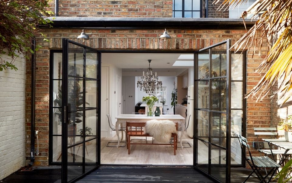 Elegant dining room viewed through large open black steel-framed glass doors, featuring a rustic table with a chandelier above and a cosy sheepskin throw on a chair, blending indoor luxury with outdoor charm in a rear home extension design.