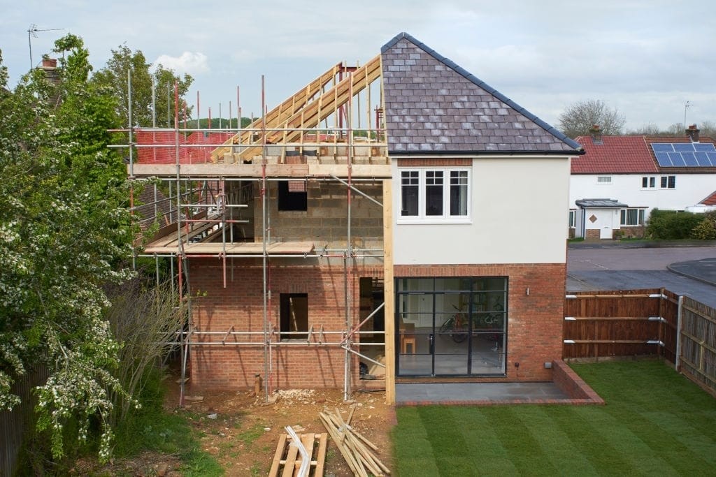 Drone photograph of the rear of a large family dwelling house which on the left hand side is currently being constructed with scaffolding still set up and roof has only the timber frame in place whilst the right hand side is the completed view of the dwelling with large bifold windows leading onto the luscious green lawn 