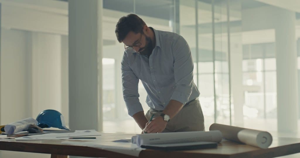 Architect in a blue shirt and beige trousers reviewing printed construction drawings on a large wooden table in the middle of the construction site whilst construction project monitoring