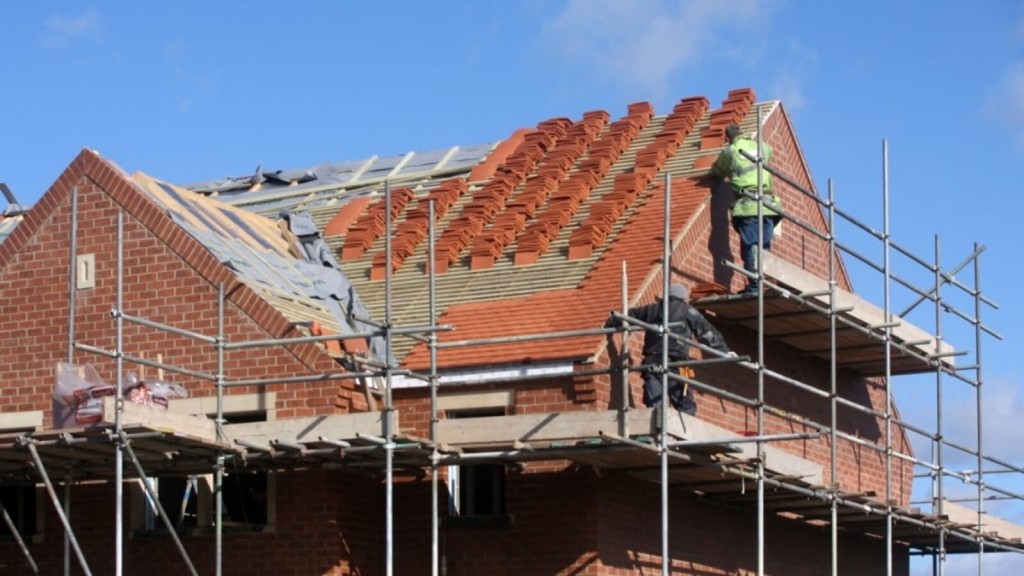 Construction worker in high-visibility jacket carefully installing red roof tiles on a new brick house, with scaffolding surrounding the structure under clear skies, showcasing skilled labor and precision in residential construction.