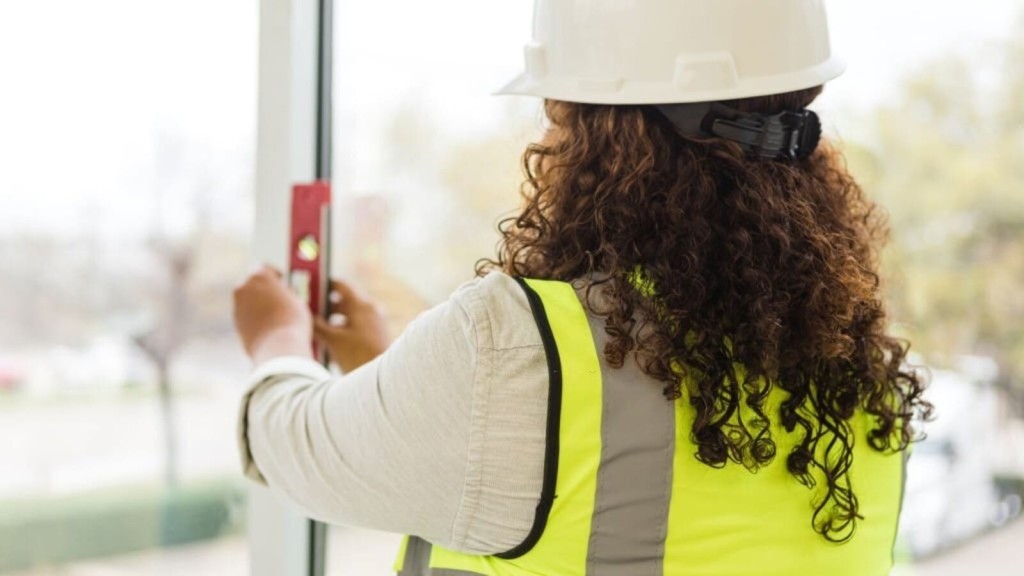 Rear view of a female construction inspector with curly hair wearing a safety helmet and reflective vest using a spirit level on a window at a construction site, ensuring quality control standards.