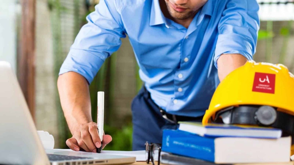 Engineer in a blue shirt focusing on architectural plans with a safety helmet, books, and drafting tools on the desk, symbolising professional project planning in construction.