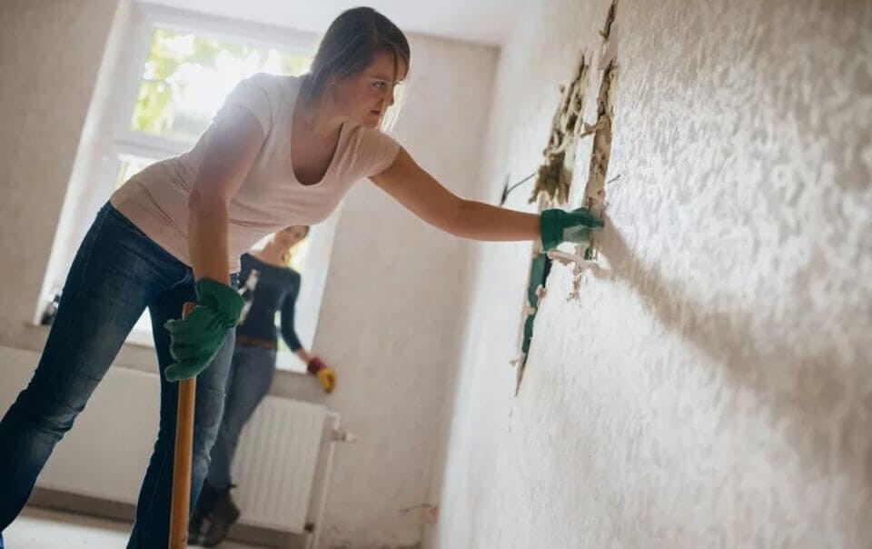 Focused woman wearing gloves removing plaster from an internal wall during a home renovation, with another person working in the background, representing DIY home improvement.