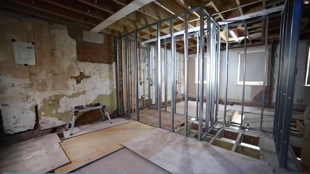 Interior construction site with metal stud framing for a new wall, exposed wooden beams, and old plaster on brickwork, indicating a work-in-progress house renovation.