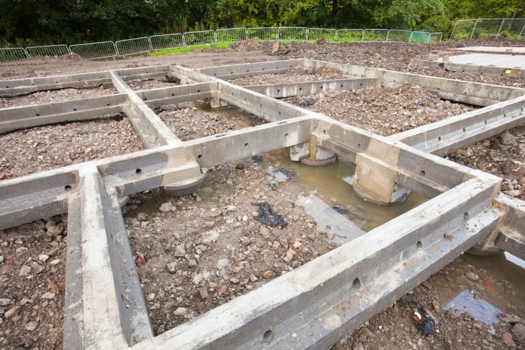 Foundation framework of a new construction site with concrete beams and footings, showing the initial stages of a building project in a muddy construction area with safety fences in the background.