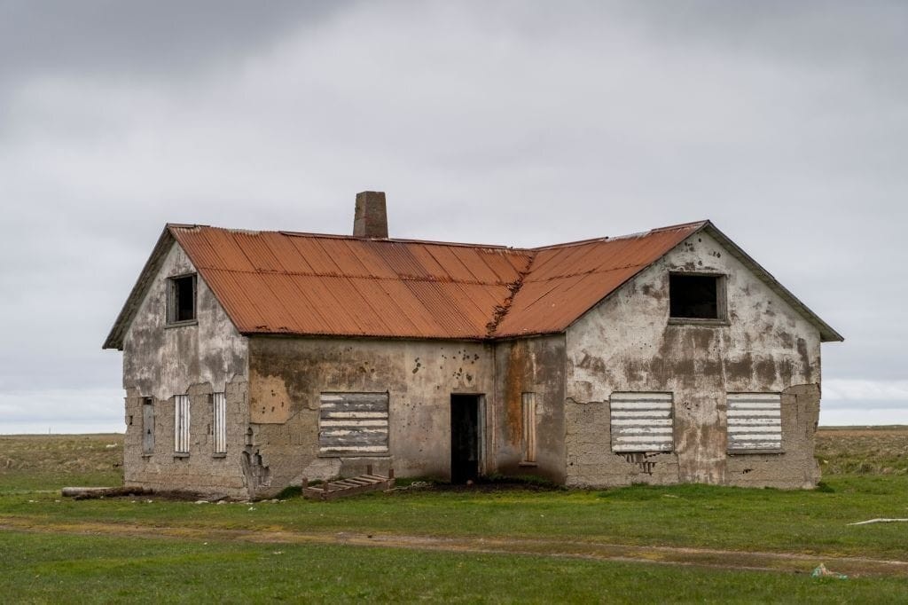 Dilapidated farmhouse with a rusted tin roof and boarded-up windows set against a grey overcast sky, symbolizing rural decay and the potential for property redevelopment.
