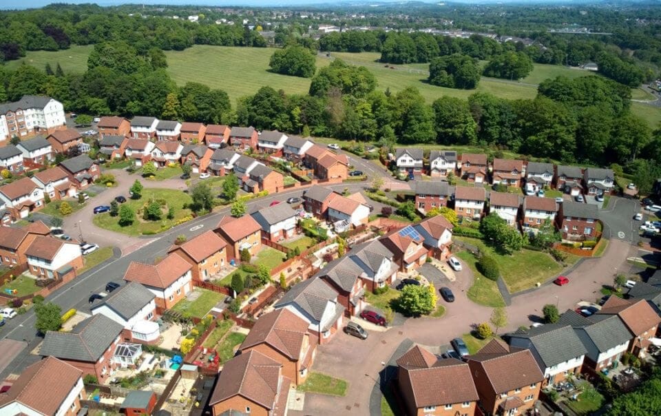 Aerial view of a residential neighbourhood with a mix of detached and semi-detached brick houses, manicured gardens, winding roads, and parked cars, adjacent to a lush green field, depicting suburban development and community planning.