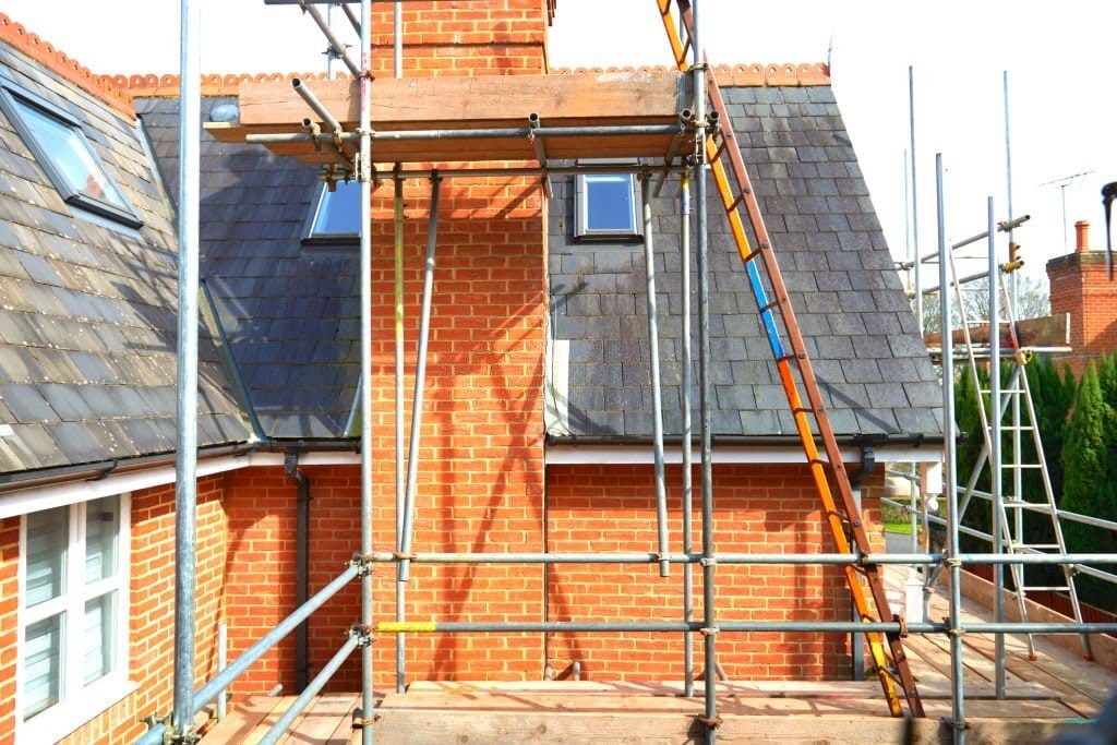 Scaffolding set up around a brick house with a slate roof in the UK, indicating ongoing construction or renovation work, with ladders for access and clear skies in the background.