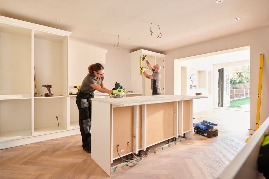 Two tradespeople installing cabinetry and shelves in a bright, modern UK kitchen during a home renovation project, with a focus on interior design and craftsmanship.