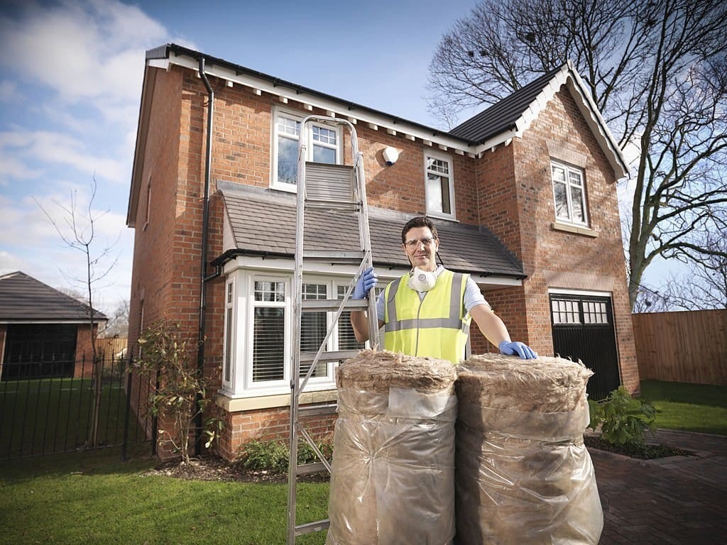 Construction professional in high-visibility vest standing in front of a UK brick detached house with insulation bags, indicating home energy retrofitting for sustainable living.