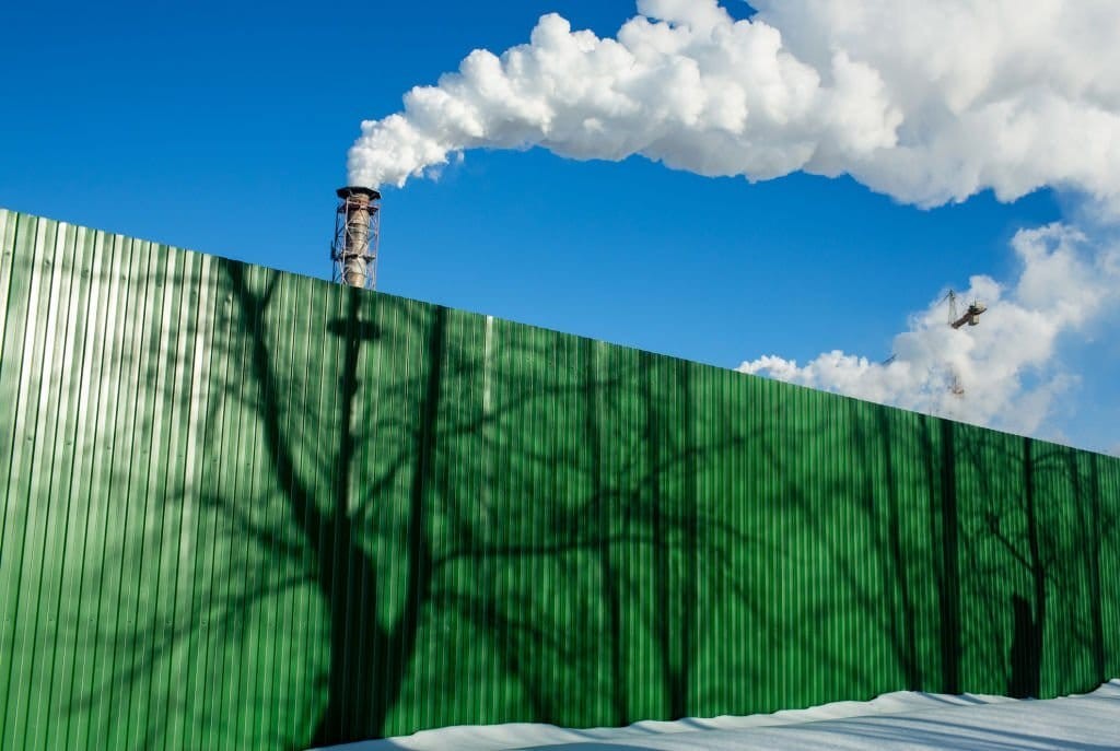 Industrial smokestack emitting steam against a clear blue sky with a green corrugated fence with casting tree shadows, and a crane in the background.