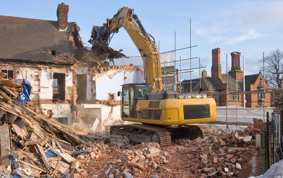 Yellow excavator demolishing an old brick house with rubble scattered around, showcasing the initial stage of a construction project for site clearance and redevelopment.