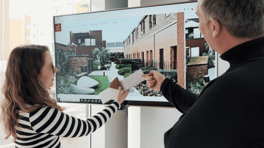 Two architects discussing exterior design plans on a digital screen in a modern office. The female architect in a striped shirt holds up material samples, while the male architect points at the screen displaying a landscaped residential development. The meeting highlights Urbanist Architecture's collaborative approach to planning variations and design decisions.